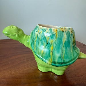 Puede incluir: Una maceta de cerámica con forma de tortuga en tonos verdes. El caparazón de la tortuga tiene un acabado texturizado y vidriado con vetas amarillas y verdes. La maceta está diseñada con una gran abertura para plantas.