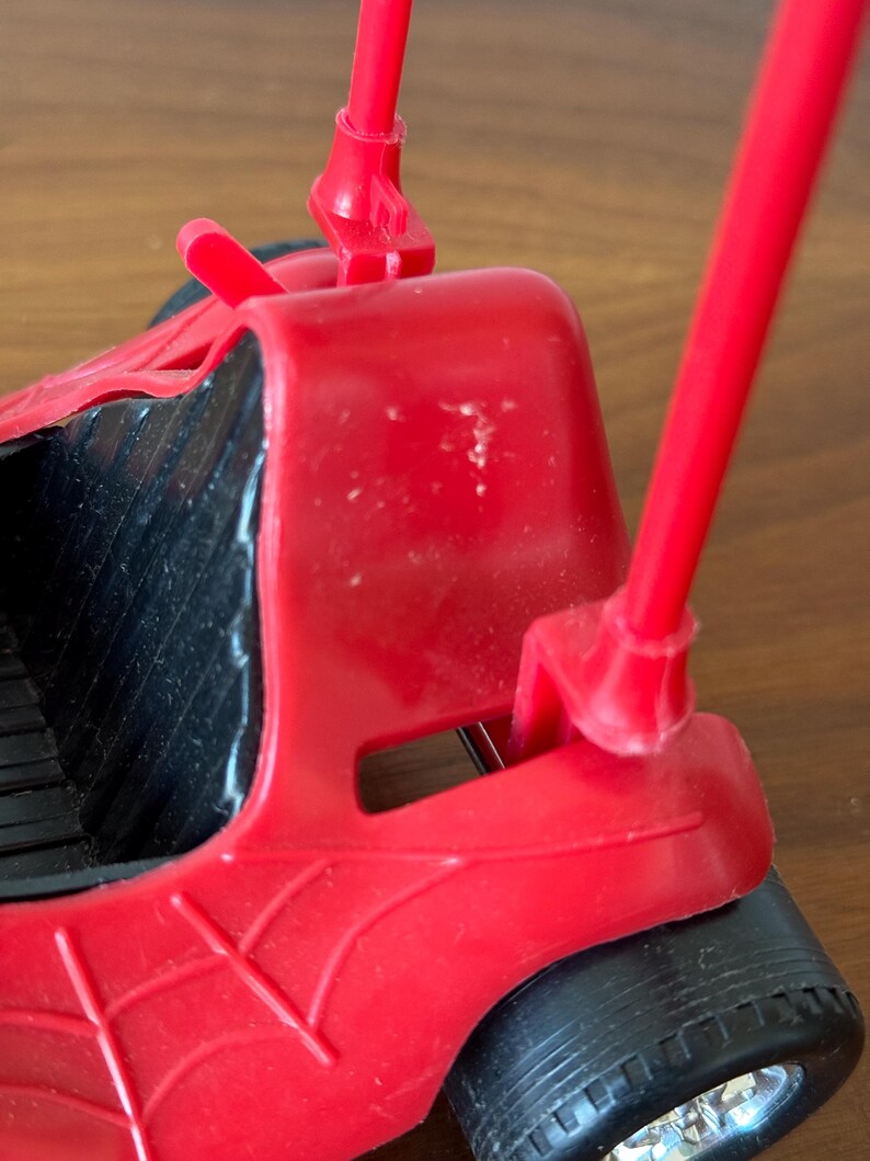 Pu&ograve; includere: Un'auto giocattolo di plastica rossa con ruote nere e un design a ragnatela. L'auto ha due pali rossi che si estendono dalla parte posteriore.