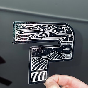 Könnte beinhalten: Ein schwarz-weißer Aufkleber in Form des Buchstabens "P" mit einem kosmischen Design. Der Aufkleber zeigt wirbelnde wolkenartige Muster, Sterne, Planeten und Bergketten. Die Ränder haben ein glitzerndes, irisierendes Finish.