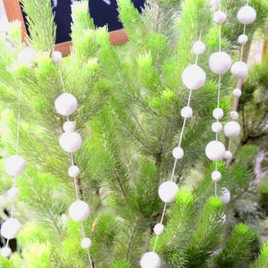 K&ouml;nnte beinhalten: Eine wei&szlig;e Pompon-Girlande h&auml;ngt an einem gr&uuml;nen Kiefernbaum. Die Girlande besteht aus wei&szlig;en Filzkugeln, die mit wei&szlig;em Garn aneinandergereiht sind. Ein Schild aus Schiefer mit dem Wort "MRS." in wei&szlig;er Kreide geschrieben, h&auml;ngt am Baum.