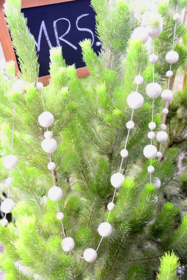 K&ouml;nnte beinhalten: Eine wei&szlig;e Pompon-Girlande h&auml;ngt an einem gr&uuml;nen Kiefernbaum. Die Girlande besteht aus wei&szlig;en Filzkugeln, die mit wei&szlig;em Garn aneinandergereiht sind. Ein Schild aus Schiefer mit dem Wort "MRS." in wei&szlig;er Kreide geschrieben, h&auml;ngt am Baum.