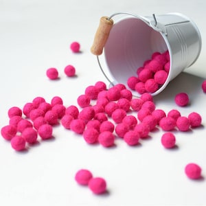 May include: A white metal bucket with a wooden handle filled with pink felt balls. The balls are spilling out of the bucket onto a white surface.