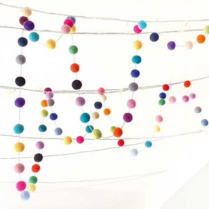 Puede incluir: Una guirnalda colorida de bolas de fieltro enhebradas en un alambre blanco. La guirnalda est&aacute; compuesta por varios colores, incluyendo rojo, naranja, amarillo, verde, azul, morado y rosa.