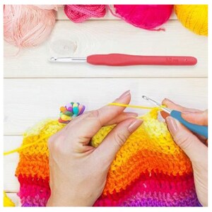 Pu&ograve; includere: Una persona sta lavorando a crochet una coperta arcobaleno colorata con un uncinetto blu. La coperta &egrave; realizzata con una variet&agrave; di colori, tra cui giallo, arancione, rosso, rosa e viola. La persona tiene l'uncinetto nella mano destra e la lana nella mano sinistra. L'uncinetto &egrave; etichettato "5.0 mm".