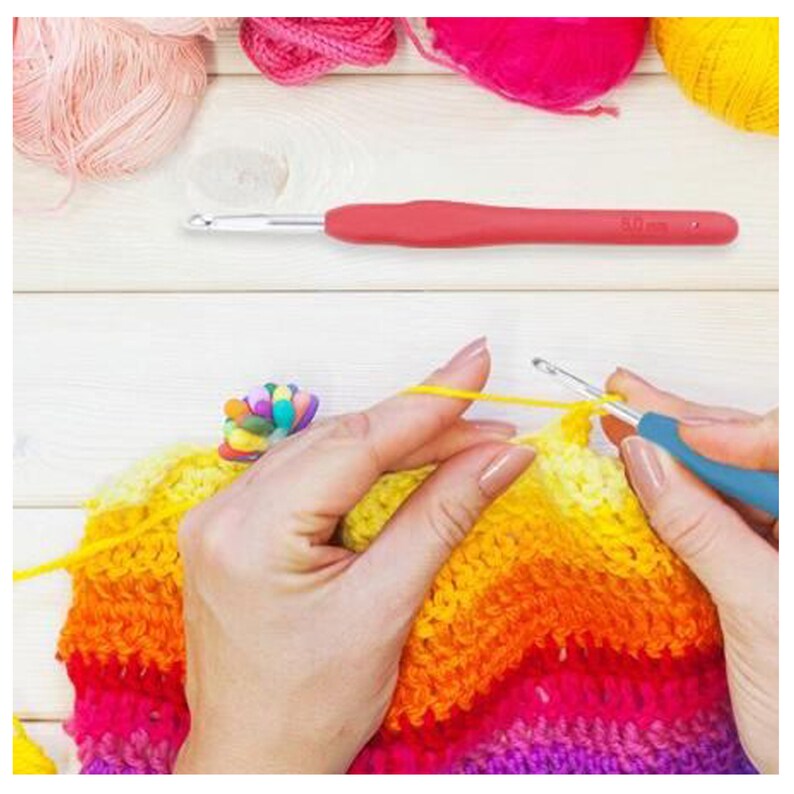 Pu&ograve; includere: Una persona sta lavorando a crochet una coperta arcobaleno colorata con un uncinetto blu. La coperta &egrave; realizzata con una variet&agrave; di colori, tra cui giallo, arancione, rosso, rosa e viola. La persona tiene l'uncinetto nella mano destra e la lana nella mano sinistra. L'uncinetto &egrave; etichettato "5.0 mm".
