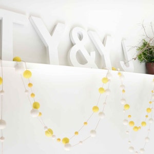 K&ouml;nnte beinhalten: Wei&szlig;e Holzbuchstaben, die "T Y & Y" buchstabieren, sind auf einem wei&szlig;en Regal angebracht. Zwei Girlanden aus gelben und wei&szlig;en Pompons h&auml;ngen vom Regal.