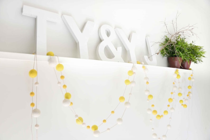 K&ouml;nnte beinhalten: Wei&szlig;e Holzbuchstaben, die "T Y & Y" buchstabieren, sind auf einem wei&szlig;en Regal angebracht. Zwei Girlanden aus gelben und wei&szlig;en Pompons h&auml;ngen vom Regal.