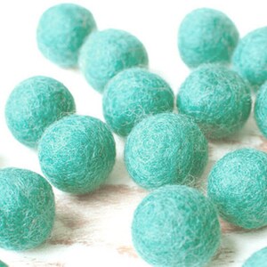 Könnte beinhalten: Eine Ansammlung von türkisfarbenen Filzkugeln, die auf einer weißen Oberfläche verstreut sind. Die Kugeln sind rund und haben eine strukturierte, wollige Erscheinung. Die Farbe ist ein lebendiger, mittlerer Türkiston, der eine spielerische und taktile Ästhetik erzeugt. Bastelbedarf.