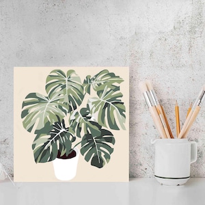 Pode incluir: Uma impressão de arte quadrada com uma ilustração estilizada de uma planta Monstera em um vaso branco sobre um fundo creme. A planta tem folhas verdes grandes e divididas. Uma caneca branca com pincéis e lápis está por perto.