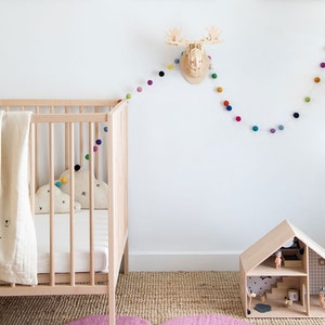 Puede incluir: Una cuna de madera blanca con una manta blanca y una almohada con forma de nube. Una casa de mu&ntilde;ecas de madera con un techo blanco y una guirnalda de pompones de colores cuelga de la pared detr&aacute;s de la cuna.