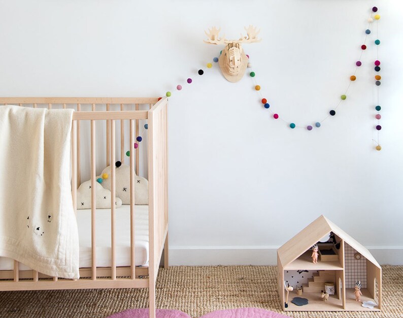 Puede incluir: Una cuna de madera blanca con una manta blanca y una almohada con forma de nube. Una casa de mu&ntilde;ecas de madera con un techo blanco y una guirnalda de pompones de colores cuelga de la pared detr&aacute;s de la cuna.