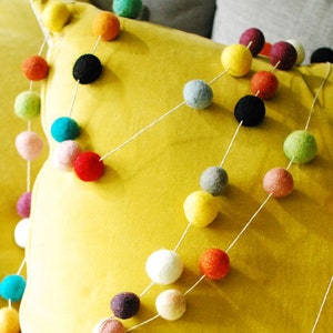 Puede incluir: Una guirnalda de bolas de fieltro de colores con una variedad de colores que incluyen rojo, verde, azul, amarillo, blanco y gris. La guirnalda est&aacute; enhebrada en una cuerda blanca y se coloca sobre una almohada amarilla.