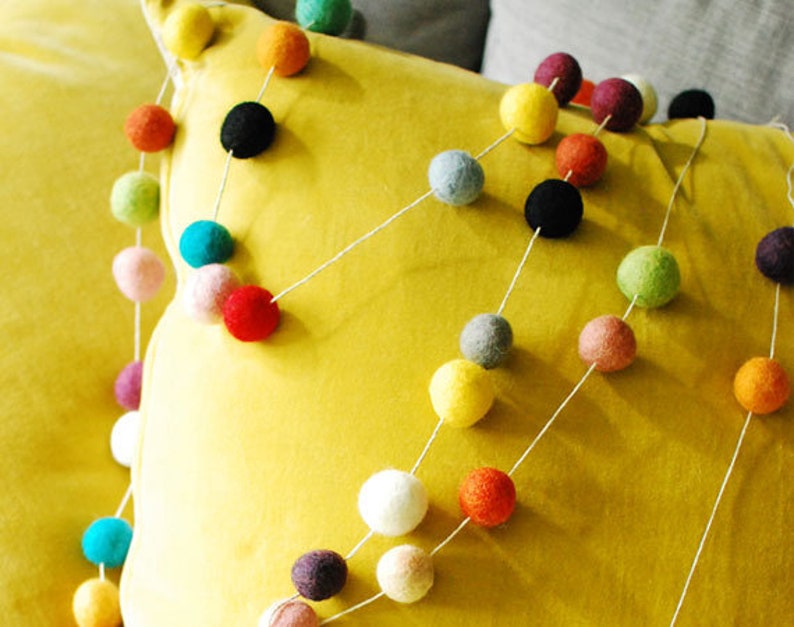 Puede incluir: Una guirnalda de bolas de fieltro de colores con una variedad de colores que incluyen rojo, verde, azul, amarillo, blanco y gris. La guirnalda est&aacute; enhebrada en una cuerda blanca y se coloca sobre una almohada amarilla.