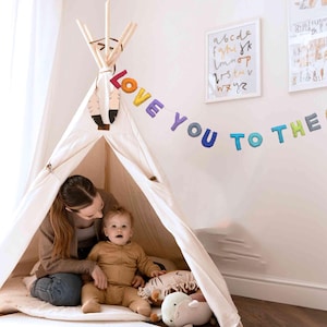 Puede incluir: Una tienda de campaña tipi blanca con una pancarta de fieltro de colores que dice "Love You To The Moon & Back" colgada encima. Una mujer y un niño están sentados dentro de la tienda de campaña.