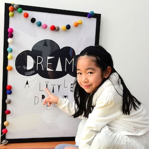Puede incluir: Un p&oacute;ster en blanco y negro con el texto "Dream a little dream" y un gr&aacute;fico de nube. Una ni&ntilde;a peque&ntilde;a est&aacute; se&ntilde;alando el p&oacute;ster, que est&aacute; decorado con una guirnalda de bolas de fieltro de colores.