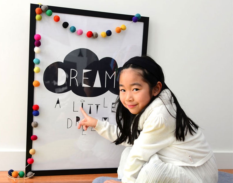 Puede incluir: Un p&oacute;ster en blanco y negro con el texto "Dream a little dream" y un gr&aacute;fico de nube. Una ni&ntilde;a peque&ntilde;a est&aacute; se&ntilde;alando el p&oacute;ster, que est&aacute; decorado con una guirnalda de bolas de fieltro de colores.