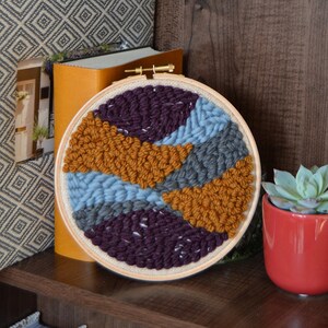 May include: A round embroidery hoop with a colorful abstract design. The design features shades of purple, blue, gray, and orange yarn. The hoop is sitting on a wooden shelf with books and a small potted succulent plant.