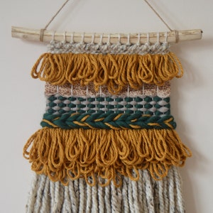 Puede incluir: Un tapiz tejido para colgar en la pared con una barra de madera natural en la parte superior. El tapiz presenta una variedad de texturas y colores, incluyendo amarillo mostaza, verde y blanco.