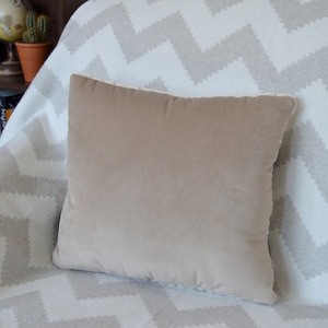 Puede incluir: Una almohada de terciopelo beige con ribete blanco se encuentra sobre una manta de tejido blanco y gris con patr&oacute;n de chevron.