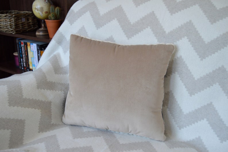 Puede incluir: Una almohada de terciopelo beige con ribete blanco se encuentra sobre una manta de tejido blanco y gris con patr&oacute;n de chevron.