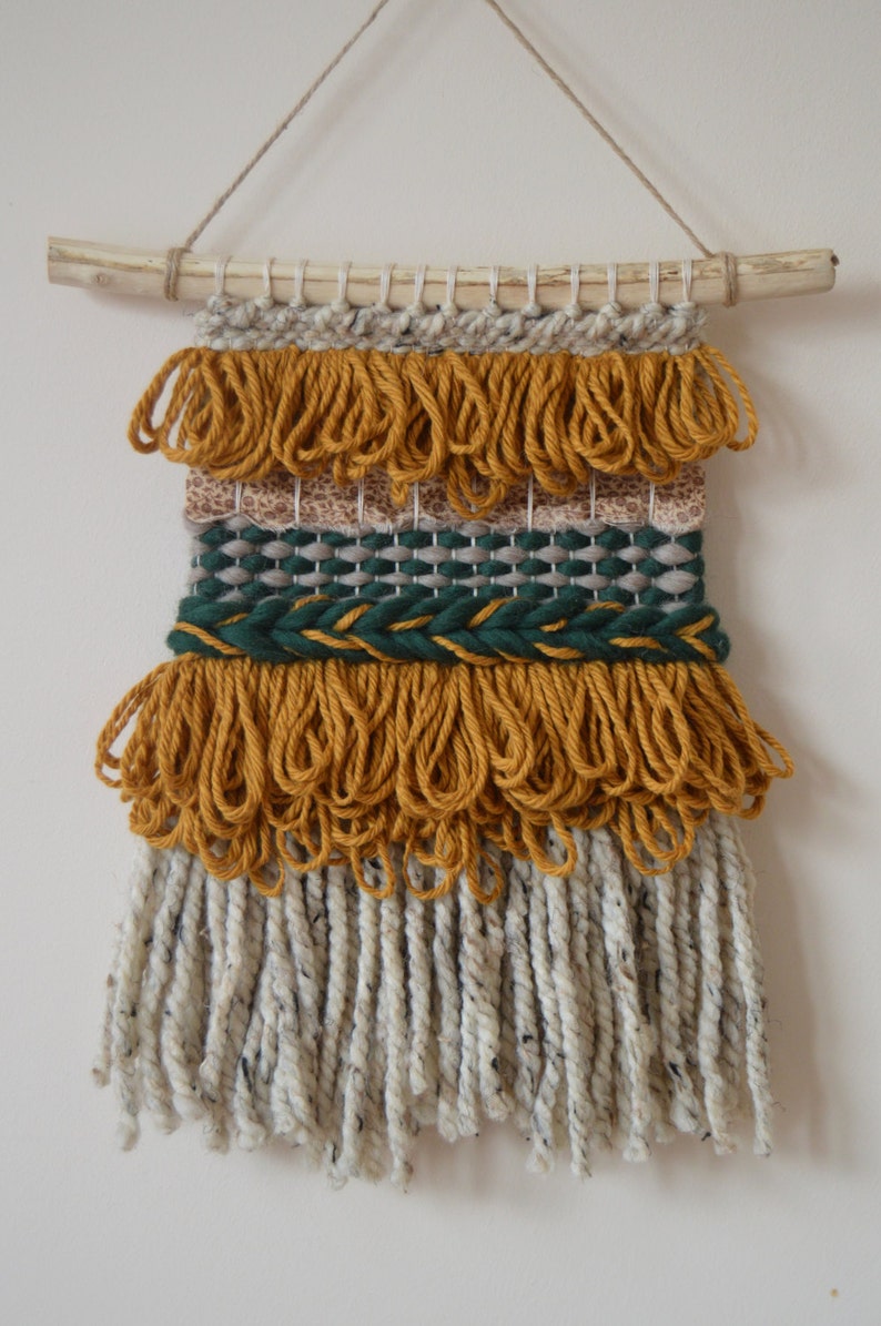 Puede incluir: Un tapiz tejido para colgar en la pared con una barra de madera natural en la parte superior. El tapiz presenta una variedad de texturas y colores, incluyendo amarillo mostaza, verde y blanco.
