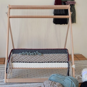 Puede incluir: Un telar de madera con un tapiz parcialmente tejido en tonos de gris, blanco y rojo. Tres ovillos de hilo est&aacute;n en primer plano, uno es gris claro, otro es azul verdoso claro y otro es negro.
