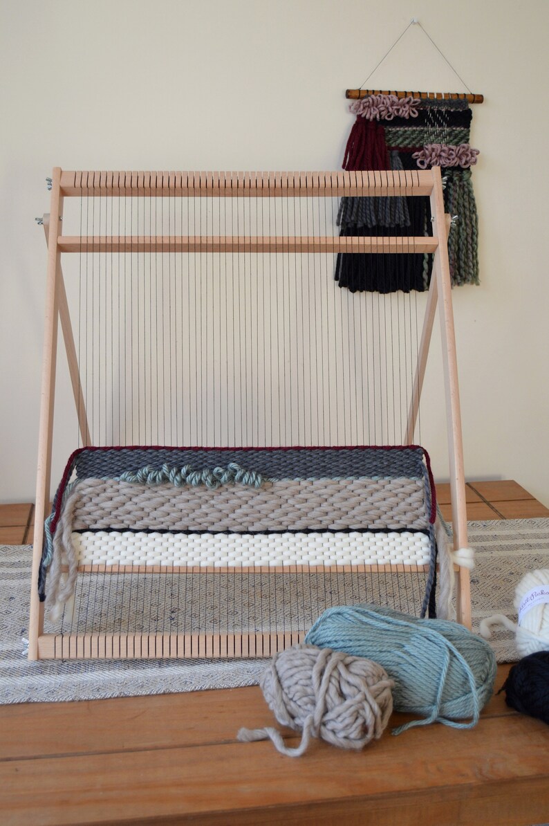 Puede incluir: Un telar de madera con un tapiz parcialmente tejido en tonos de gris, blanco y rojo. Tres ovillos de hilo est&aacute;n en primer plano, uno es gris claro, otro es azul verdoso claro y otro es negro.