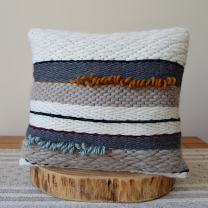 Puede incluir: Una almohada tejida con un patr&oacute;n a rayas en tonos de blanco, gris, beige y azul. La almohada tiene una superficie texturizada y presenta detalles de flecos en naranja y azul.