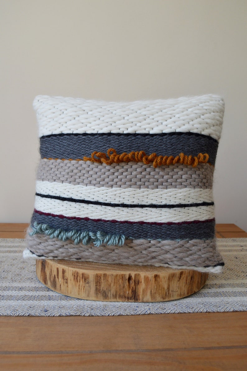 Puede incluir: Una almohada tejida con un patr&oacute;n a rayas en tonos de blanco, gris, beige y azul. La almohada tiene una superficie texturizada y presenta detalles de flecos en naranja y azul.