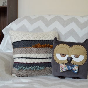 Puede incluir: Dos almohadas decorativas en un sof&aacute; con estampado de chevron blanco y gris. Una almohada est&aacute; tejida con rayas marrones, blancas, grises y verdes. La otra almohada es un b&uacute;ho marr&oacute;n con una pajarita azul y blanca.
