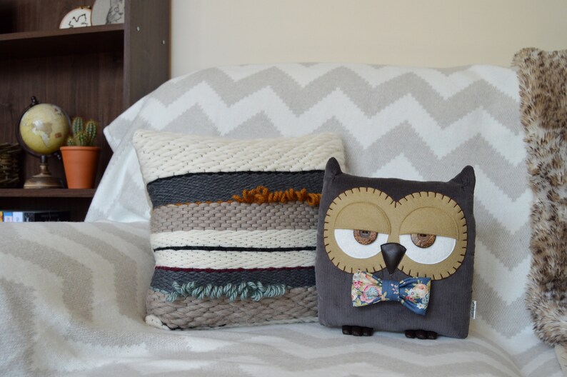 Puede incluir: Dos almohadas decorativas en un sof&aacute; con estampado de chevron blanco y gris. Una almohada est&aacute; tejida con rayas marrones, blancas, grises y verdes. La otra almohada es un b&uacute;ho marr&oacute;n con una pajarita azul y blanca.