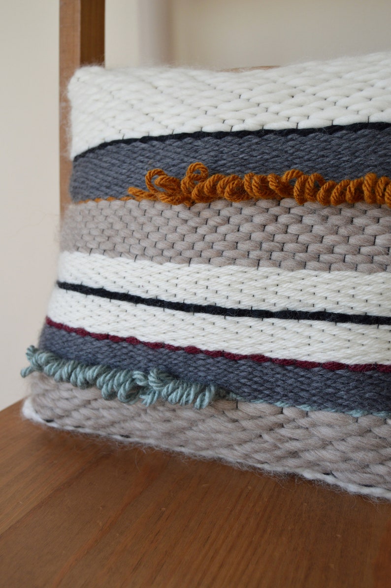 Puede incluir: Una almohada tejida con un patr&oacute;n a rayas en tonos de blanco, gris, marr&oacute;n y rojo. La almohada tiene un detalle de flecos en naranja, verde y gris.