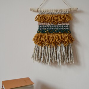 Puede incluir: Un tapiz de pared hecho de hilo tejido con una varilla de madera natural en la parte superior. El hilo es en tonos de amarillo mostaza, beige y verde bosque. El tapiz tiene una franja de hilo blanco y amarillo mostaza.