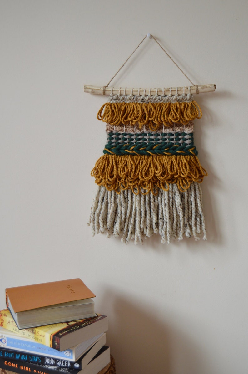 Puede incluir: Un tapiz de pared hecho de hilo tejido con una varilla de madera natural en la parte superior. El hilo es en tonos de amarillo mostaza, beige y verde bosque. El tapiz tiene una franja de hilo blanco y amarillo mostaza.
