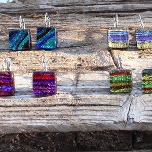 May include: A collection of square dichroic glass earrings with silver-colored ear wires. The earrings feature various colors, including blue, green, red, purple, and gold, with striped and textured patterns. The earrings are displayed on a weathered wooden surface.