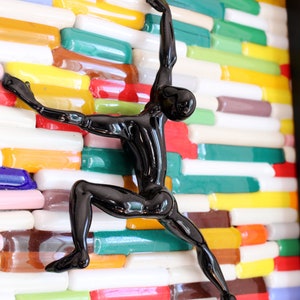 K&ouml;nnte beinhalten: Eine schwarze Glasskulptur einer Person, die eine Wand aus bunten Glasfliesen erklimmt.