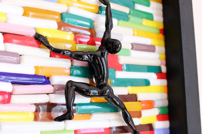 K&ouml;nnte beinhalten: Eine schwarze Glasskulptur einer Person, die eine Wand aus bunten Glasfliesen erklimmt.