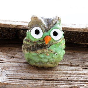 May include: A green glass owl figurine with large white eyes and an orange beak. The owl is sitting on a piece of weathered wood.