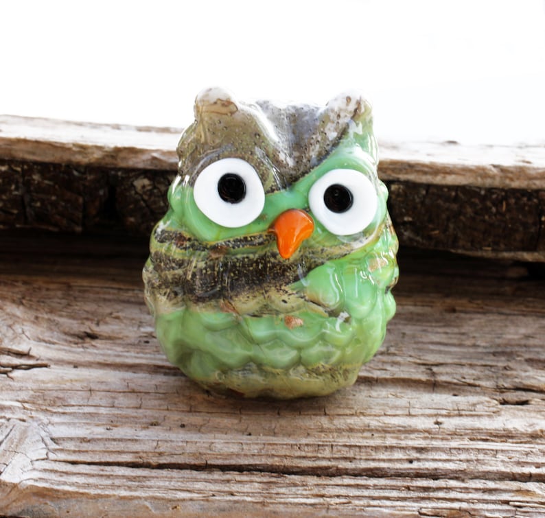 May include: A green glass owl figurine with large white eyes and an orange beak. The owl is sitting on a piece of weathered wood.
