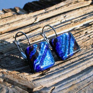 May include: A pair of square earrings with a dichroic glass design. The earrings feature a vibrant blue and black color scheme with silver-tone lever-back closures. The earrings are displayed on a weathered wooden surface.