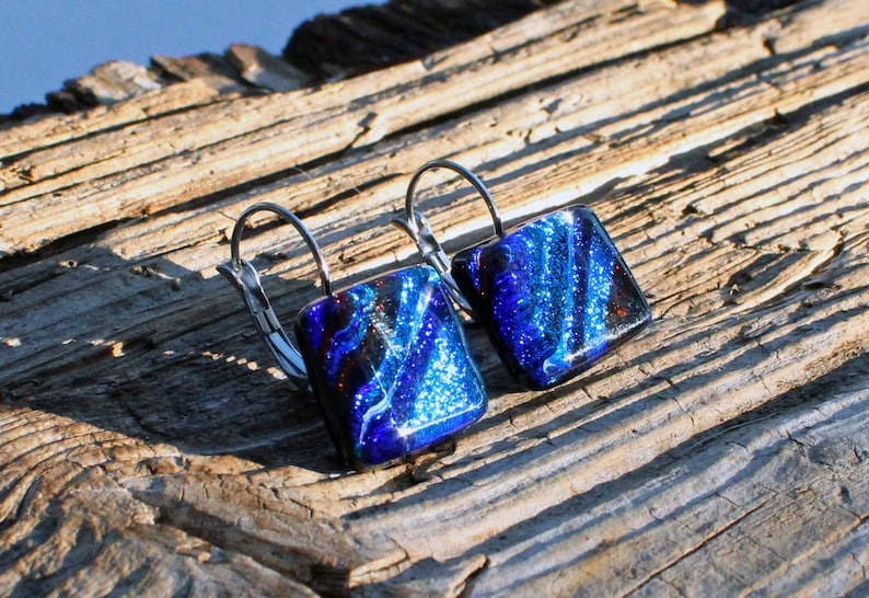May include: A pair of square earrings with a dichroic glass design. The earrings feature a vibrant blue and black color scheme with silver-tone lever-back closures. The earrings are displayed on a weathered wooden surface.