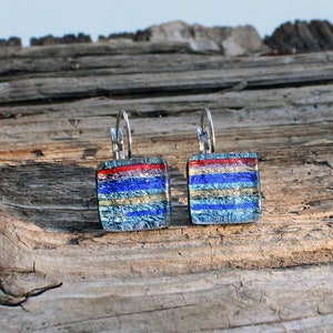 May include: Square dichroic glass earrings with a layered design. The earrings feature a red stripe at the top, followed by blue, gold, and silver stripes. The earrings have silver lever-back closures.
