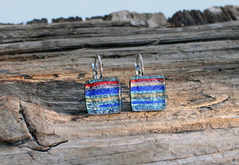 May include: Square dichroic glass earrings with a layered design. The earrings feature a red stripe at the top, followed by blue, gold, and silver stripes. The earrings have silver lever-back closures.