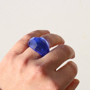 May include: A blue glass ring with a faceted design. The ring is worn on a person's finger.