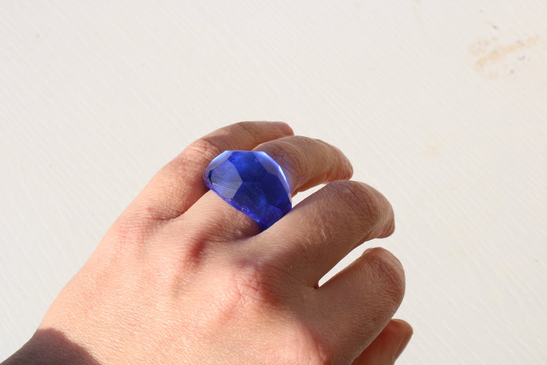 May include: A blue glass ring with a faceted design. The ring is worn on a person's finger.
