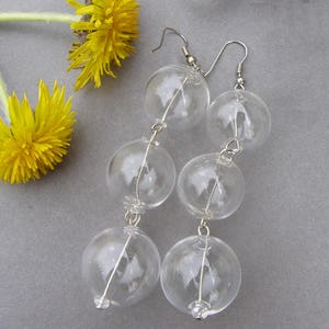 May include: Clear glass ball earrings with silver-tone hooks and wire detailing. The earrings are composed of three graduated spheres. A yellow dandelion flower is in the background.