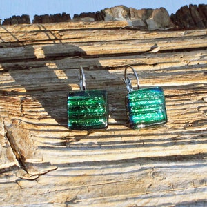 May include: A pair of square, dichroic glass earrings with a lever back closure. The earrings feature a vibrant green color with horizontal lines and a shimmering effect. The earrings are displayed on a weathered wooden surface.