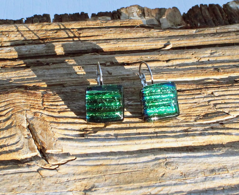 May include: A pair of square, dichroic glass earrings with a lever back closure. The earrings feature a vibrant green color with horizontal lines and a shimmering effect. The earrings are displayed on a weathered wooden surface.