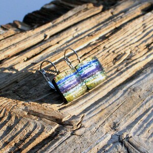 May include: Dichroic glass earrings with silver-colored leverback closures. The earrings feature a rectangular shape with layers of blue, green, and purple hues. The earrings are set against a weathered wood background.