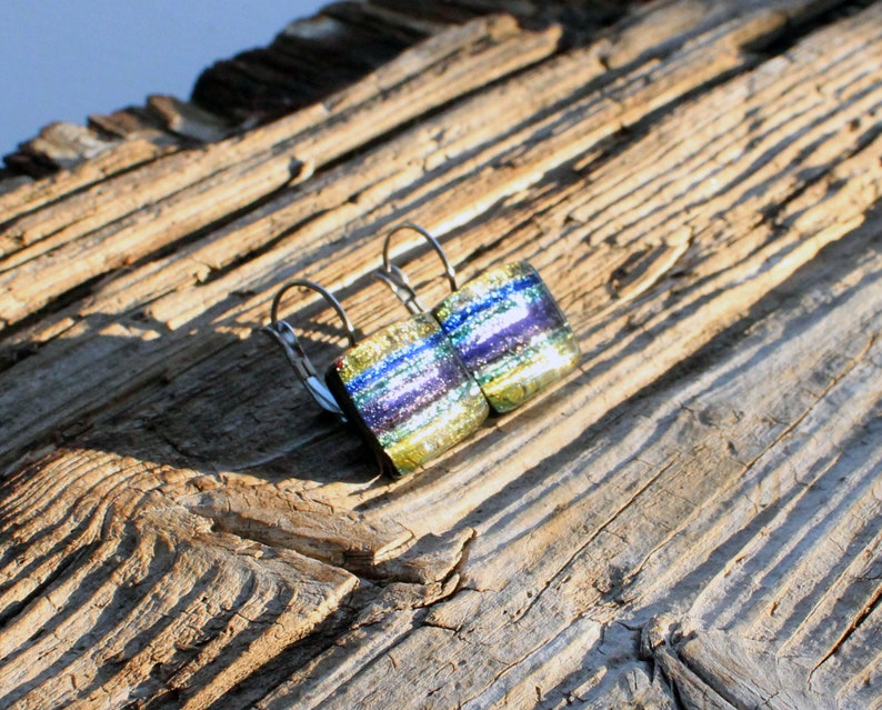 May include: Dichroic glass earrings with silver-colored leverback closures. The earrings feature a rectangular shape with layers of blue, green, and purple hues. The earrings are set against a weathered wood background.